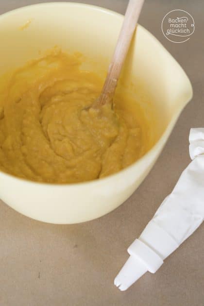 Gelber Brandteig in Rührschüssel mit Kochlöffel neben gefülltem Spritzbeutel