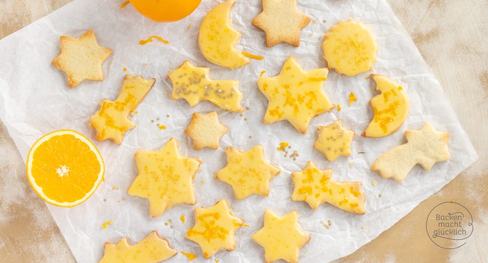 Sternförmige Orangenplätzchen mit gelbem Zuckerguss neben halbierter Orange auf Backpapier