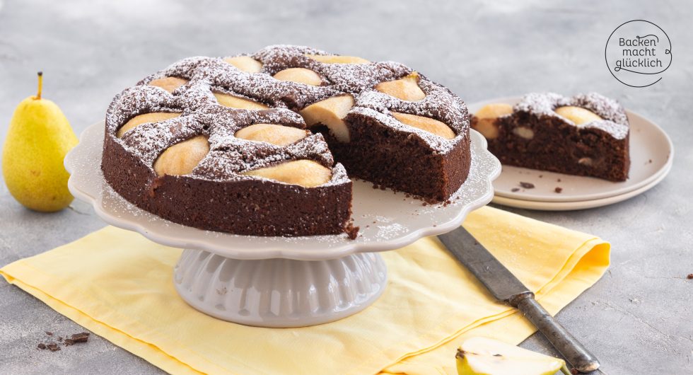 Angeschnittener Schoko-Birnenkuchen mit Puderzucker auf weißer Kuchenplatte, daneben ganze Birne