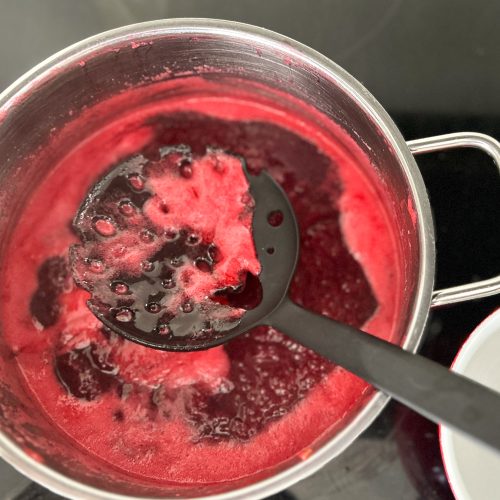 Kochende Johannisbeermarmelade im Topf mit schwarzer Schaumkelle verrührt