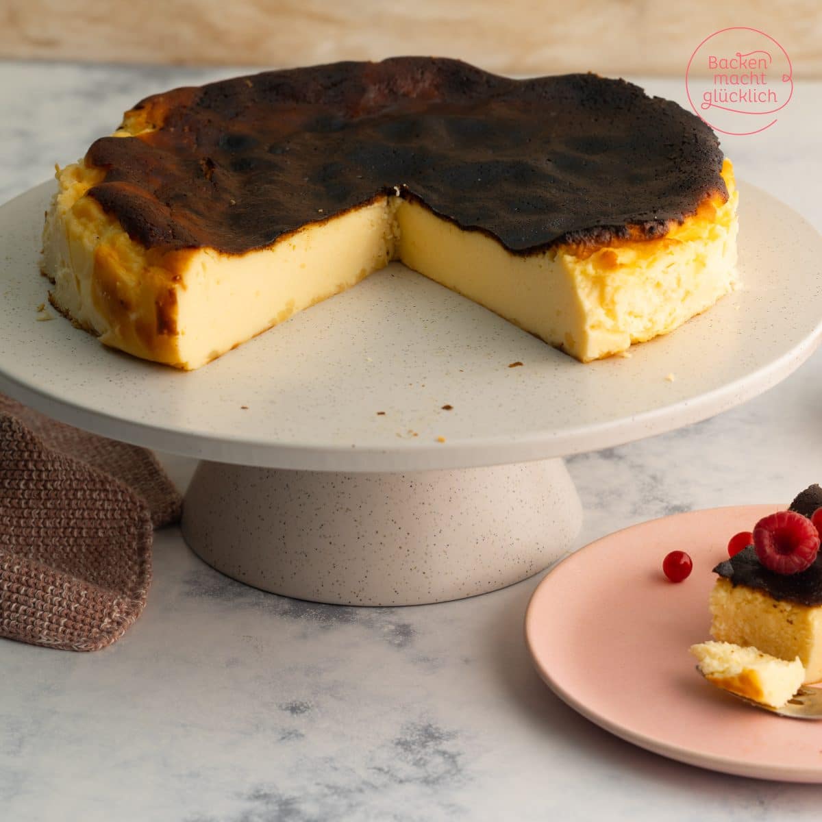 Angeschnittener San Sebastian Cheesecake mit karamellisierter Kruste auf Kuchenständer, Stück auf rosa Teller